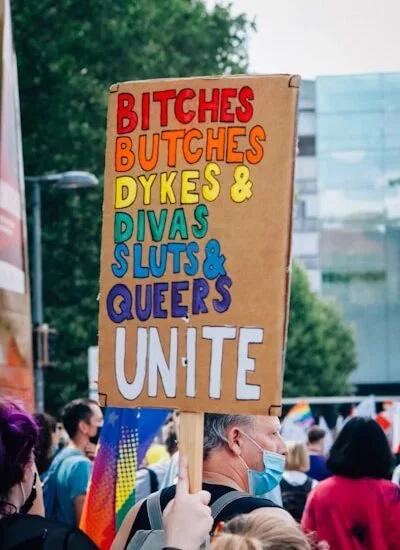 Protestplakat mit den Worten 'Bitches, Butches, Dykes, Sluts & Queers Unite' in bunten Buchstaben.